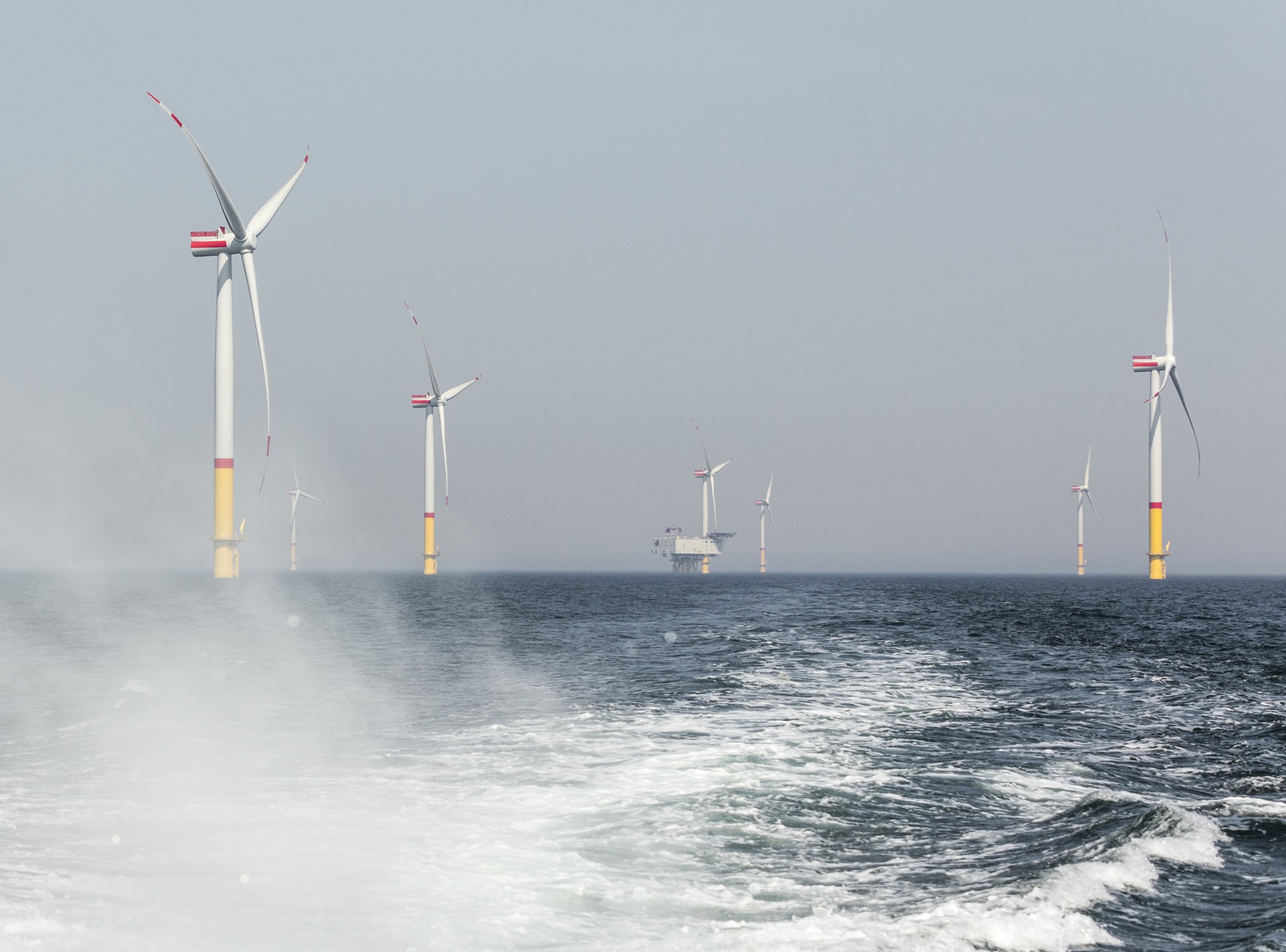A view of offshore wind turbines in a hazy sea, with choppy waters and a distant platform.