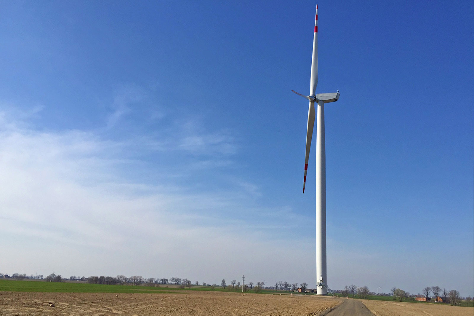 A tall wind turbine stands in a field under a bright blue sky, surrounded by farmland.