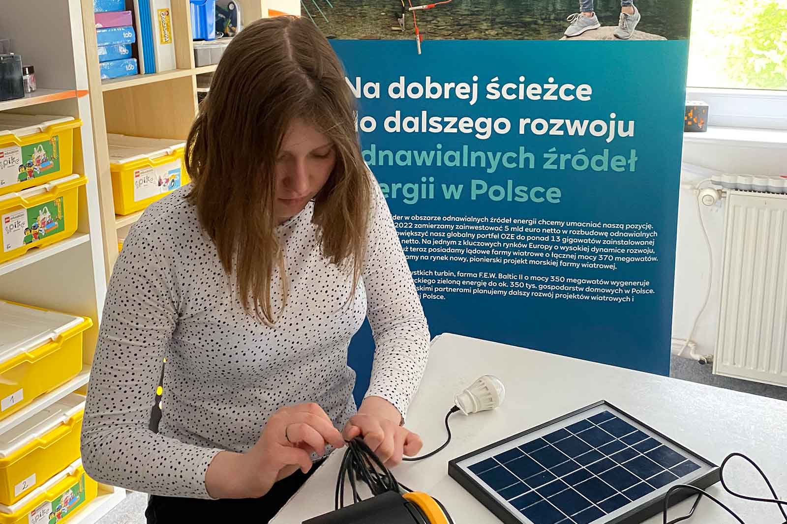 Dziewczyna pracuje z panelami słonecznymi i kablami przy stole, w tle widoczny jest banner informacyjny.