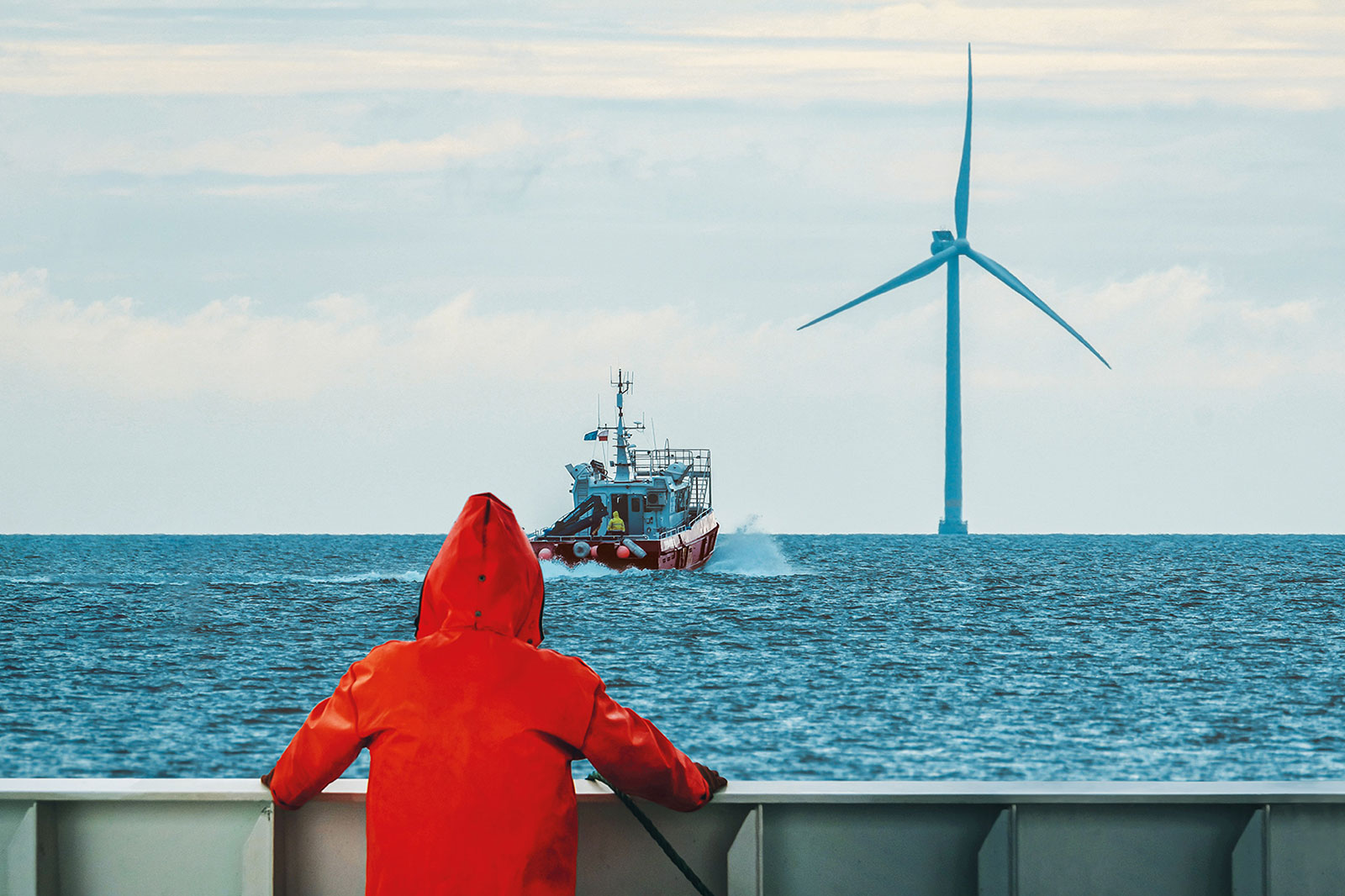 Osoba w czerwonej wodoodpornej kurtce obserwuje łódź w pobliżu turbiny wiatrowej na morzu.