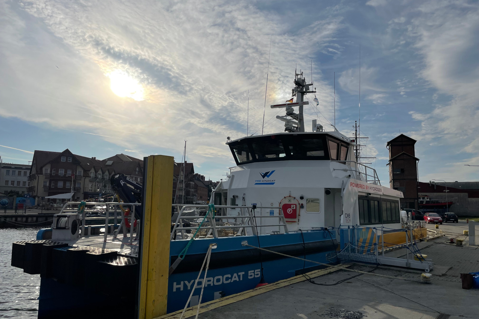 Katamaran na wodór o nazwie Hydrocat 55 zacumowany w marinie w Ustce pod pochmurnym niebem. Widać pobliskie budynki i wieżę.