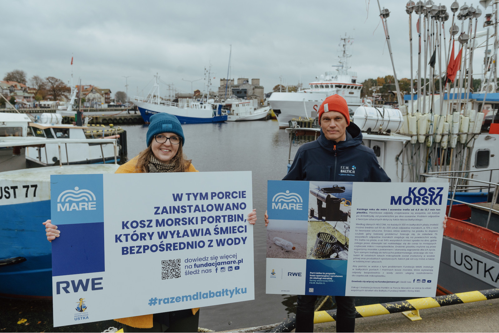 Dwie osoby trzymają znaki dotyczące inicjatywy zbierania odpadów morskich w ruchliwym porcie Ustka z różnymi łodziami w tle.