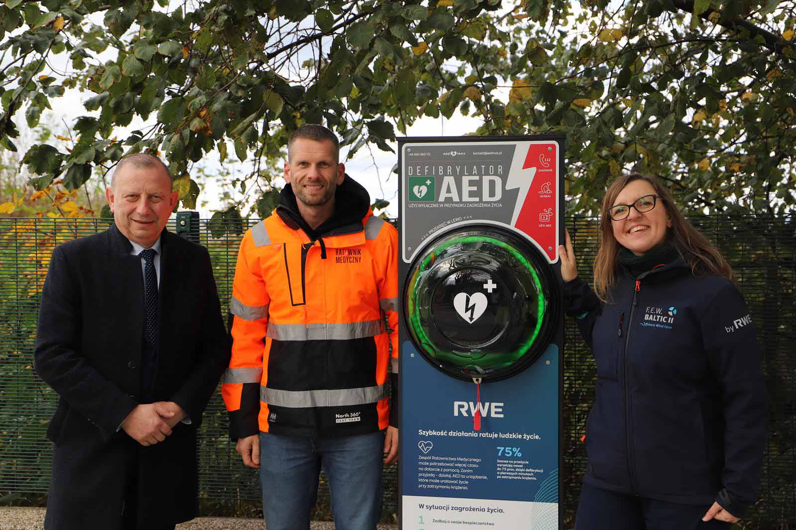 Three individuals stand next to an AED defibrillator installation in a park, surrounded by green leaves.