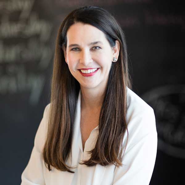 A person with long, straight hair is wearing a white shirt. Blurred writing is visible in the background, portrait of Anja-Isabel Dotzenrath.