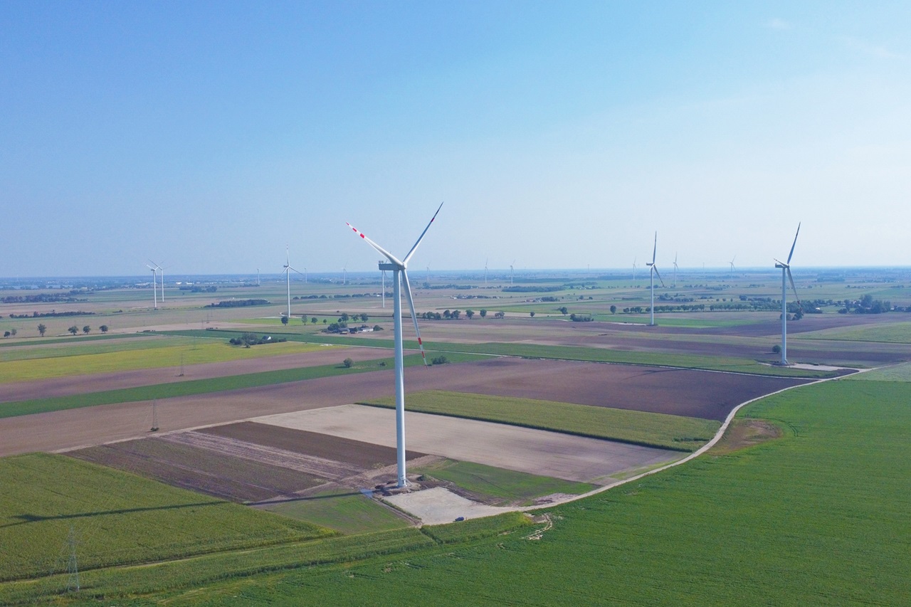 A wide landscape featuring wind turbines set against green fields and farmland.