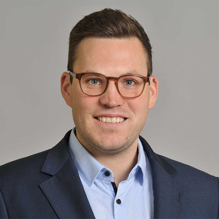 A man wearing a dark suit and a striped shirt stands against a grey background. Portrait of Benjamin Schütz.
