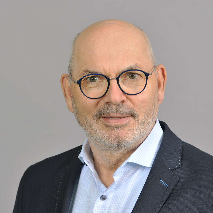 A man in a dark jacket and light blue shirt stands against a grey background. Portrait of Lothar Lambertz.