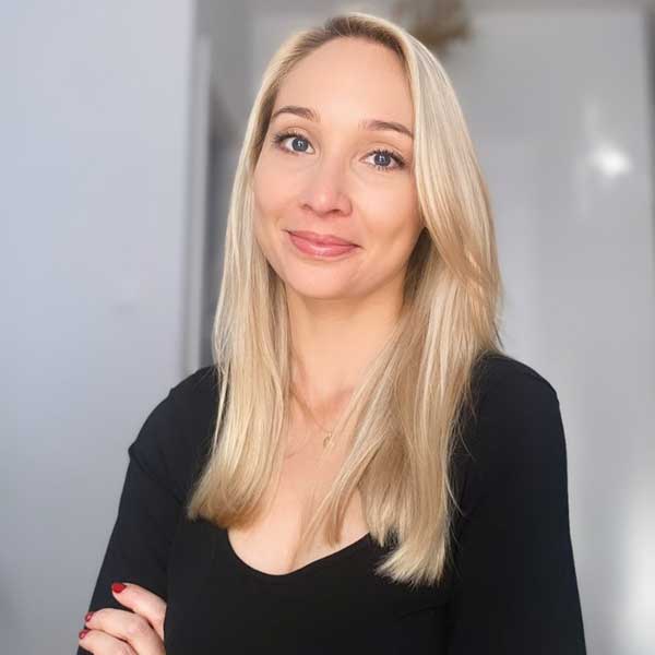 A woman with long blonde hair in a black top, standing in a bright room with a neutral background.